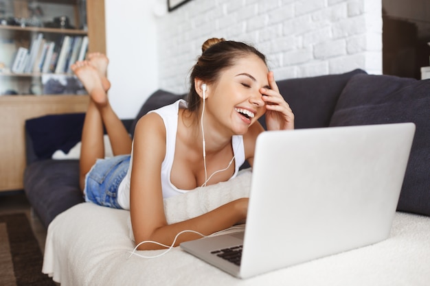 Chica joven de risa atractiva que pone en el sofá en la sala de estar con la computadora portátil.