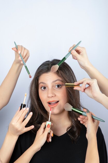Chica joven rodeada de manos de maquilladores con pinceles, lápiz labial y rímel cerca de su rostro.