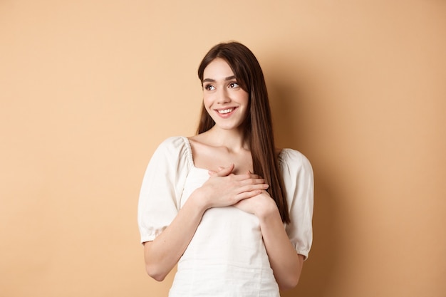 Chica joven romántica en vestido tomados de la mano en el corazón, sonriendo y mirando el espacio vacío agradecido, sintiendo gratitud, de pie sobre fondo beige