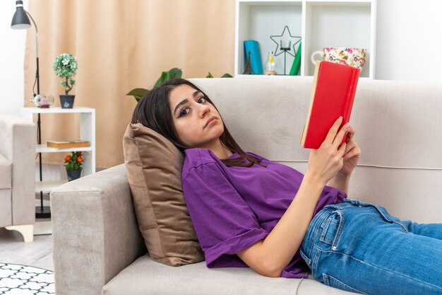 Chica joven en ropa casual con libro con expresión triste acostado en un sofá en la sala de luz