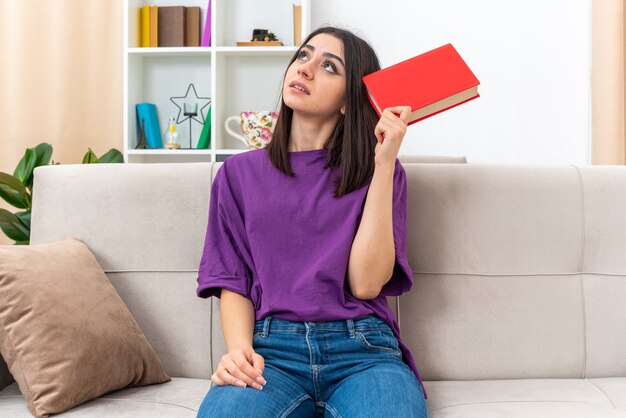 Chica joven en ropa casual con libro mirando hacia arriba desconcertado sentado en un sofá en la sala de luz