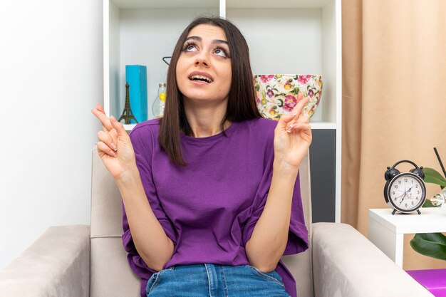 Chica joven en ropa casual mirando hacia arriba cruzando los dedos pidiendo deseo sentado en una silla en la sala de luz