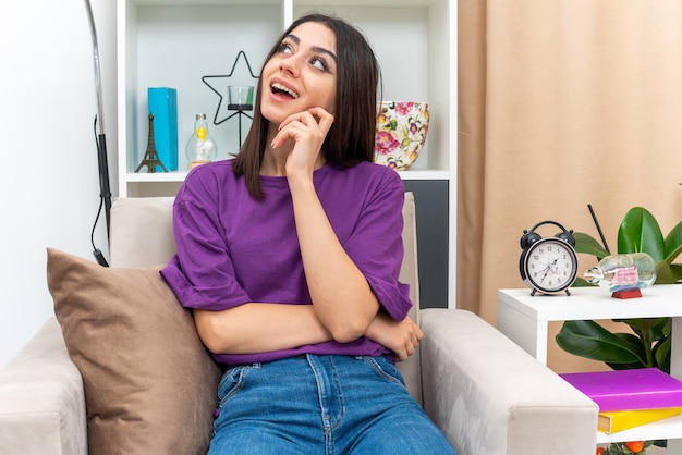 Chica joven en ropa casual mirando hacia arriba con expresión pensativa en la cara pensando en positivo sentado en una silla en la sala de luz