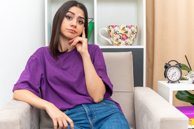 Chica joven en ropa casual mirando confundido y muy ansioso sentado en una silla en la sala de luz