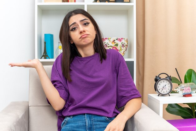 Chica joven en ropa casual mirando con expresión triste en la cara presentando algo con el brazo de su mano sentado en una silla en la sala de luz