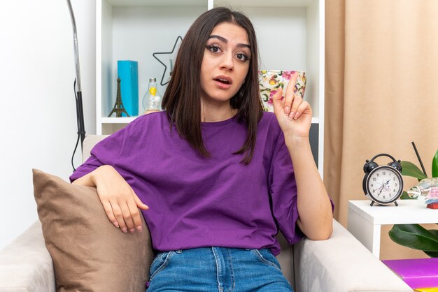 Chica joven en ropa casual mirando feliz y sorprendido mostrando el dedo índice con nueva idea sentado en una silla en la sala de luz