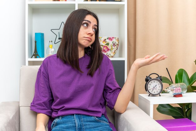 Chica joven en ropa casual mirando a un lado con expresión triste que presenta algo con el brazo de su mano sentado en un sofá en la sala de luz