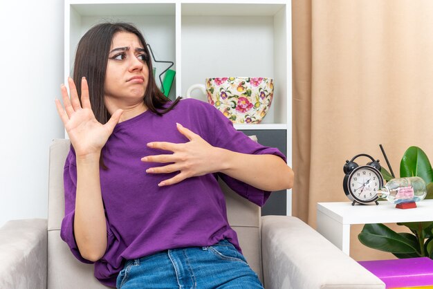 Chica joven en ropa casual mirando a un lado preocupado gesticulando con las manos sentado en una silla en la sala de luz