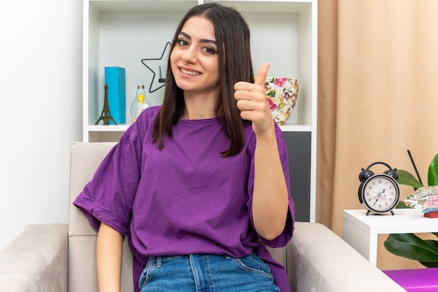 Chica joven en ropa casual mirando sonriendo alegremente feliz y positivo mostrando los pulgares para arriba sentado en una silla en la sala de luz