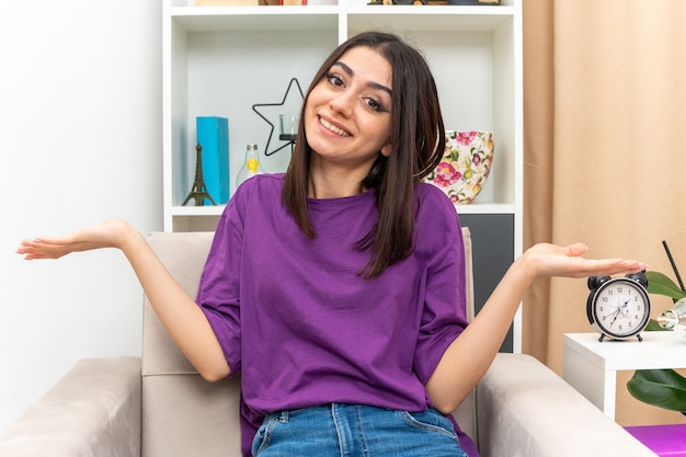 Foto gratuita chica joven en ropa casual mirando sonriendo confundido extendiendo los brazos a los lados sentado en una silla en la sala de luz