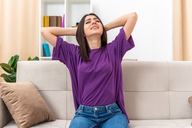 Chica joven en ropa casual que parece molesta e irritada con las manos en la cabeza sentada en un sofá en la sala de luz