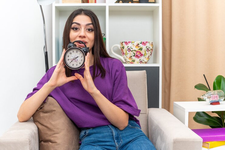 Foto gratuito chica joven en ropa casual con reloj despertador feliz y sorprendido sonriendo alegremente sentado en una silla en la sala de luz