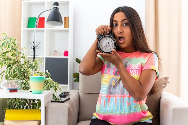 Chica joven en ropa casual con reloj despertador mirando a la cámara preocupado sentado en la silla en la sala de luz