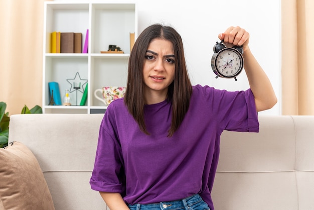 Foto gratuita chica joven en ropa casual con reloj despertador mirando confundido y disgustado sentado en un sofá en la sala de luz