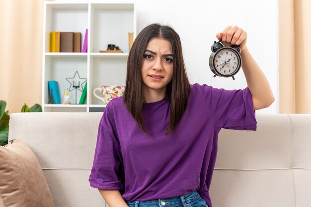 Chica joven en ropa casual con reloj despertador mirando confundido y disgustado sentado en un sofá en la sala de luz