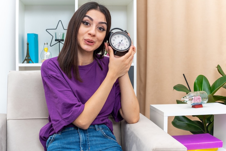 Foto gratuito chica joven en ropa casual con reloj despertador mirando con una sonrisa en la cara sentado en una silla en la sala de luz
