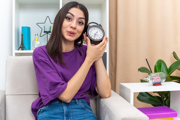 Chica joven en ropa casual con reloj despertador mirando con una sonrisa en la cara sentado en una silla en la sala de luz