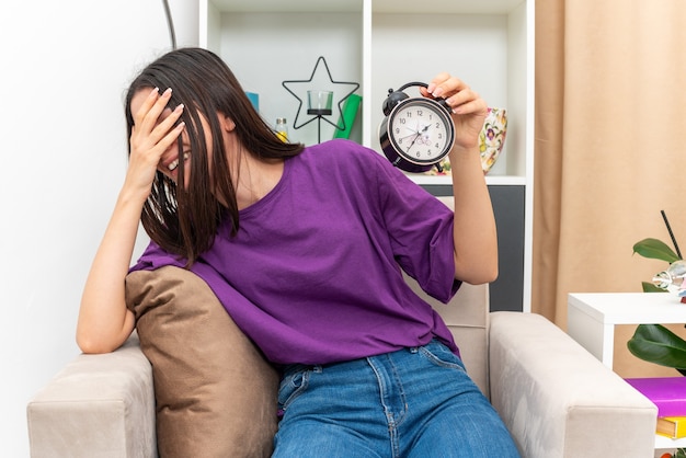 Foto gratuita chica joven en ropa casual con reloj despertador sonriendo alegremente cubriendo la cara con la palma sentada en una silla en la sala de luz