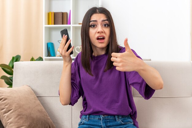 Chica joven en ropa casual con smartphone mirando a la cámara emocional y confundida mostrando los pulgares para arriba sentado en un sofá en la sala de luz