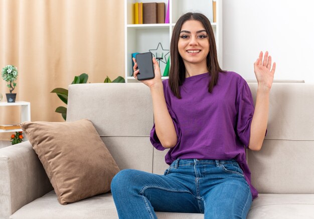 Chica joven en ropa casual con smartphone mirando a cámara sonriendo feliz y positivo saludando con la mano sentado en un sofá en la sala de luz