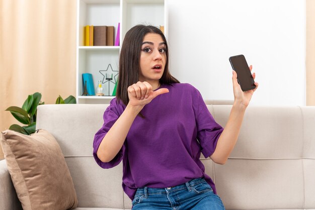 Chica joven en ropa casual con smartphone mirando confundido apuntando con el pulgar al móvil sentado en un sofá en la sala de luz