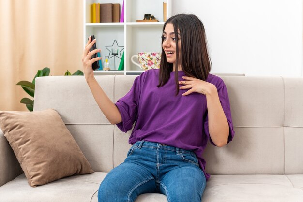 Chica joven en ropa casual con smartphone con videollamada feliz y positiva sonriendo confiada sentada en un sofá en la sala de estar luminosa