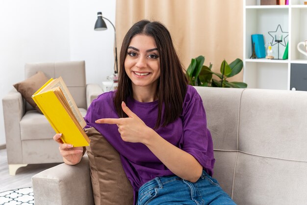 Chica joven en ropa casual sosteniendo un libro apuntando con el dedo índice a él feliz y positivo sonriendo alegremente sentado en un sofá en la sala de estar luminosa