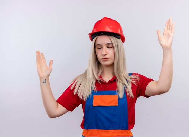 Chica joven rubia ingeniero constructor en uniforme con las manos levantadas y los ojos cerrados en la pared blanca aislada