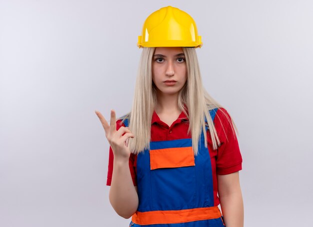 Chica joven rubia ingeniero constructor en uniforme mostrando dos mirando en la pared blanca aislada con espacio de copia