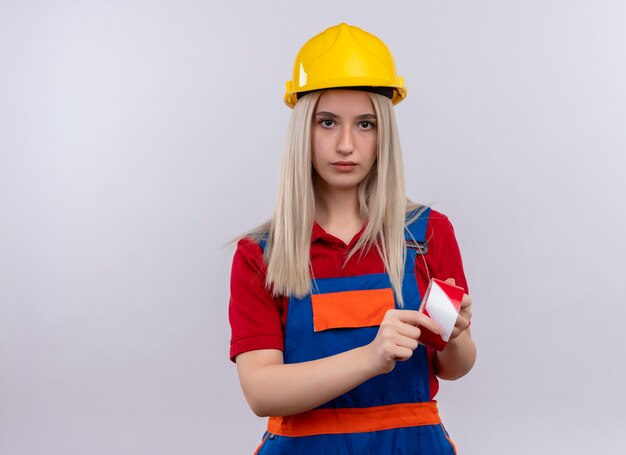 Chica joven rubia ingeniero constructor en uniforme sosteniendo cinta adhesiva mirando en la pared blanca aislada con espacio de copia