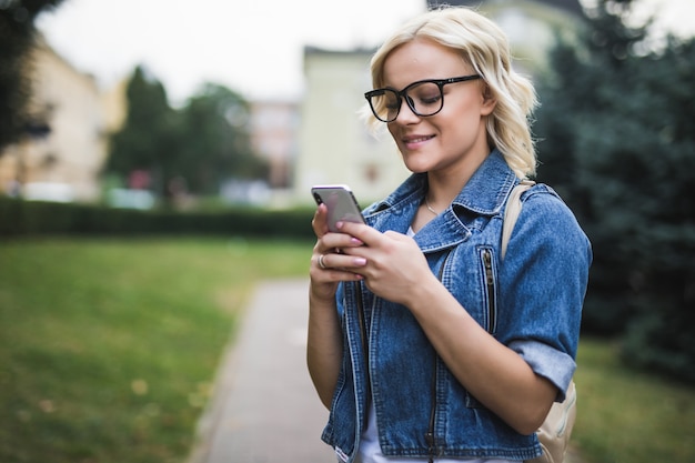 Chica joven rubia usa el teléfono para desplazarse por la conversación de la red social en la mañana de la plaza del otoño de la ciudad