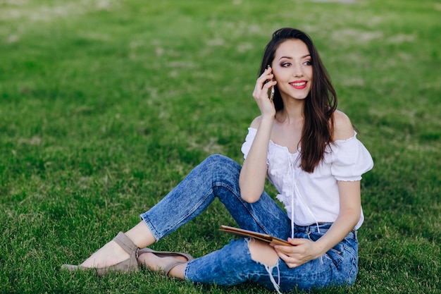 Chica joven sentada en un parque hablando por un móvil mientras sujeta una tablet