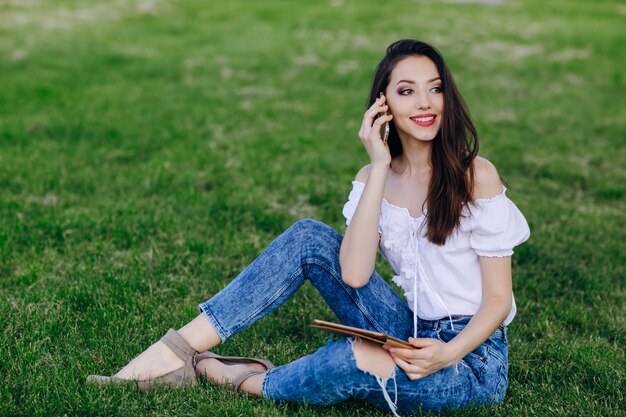 Chica joven sentada en un parque hablando por un móvil mientras sujeta una tablet