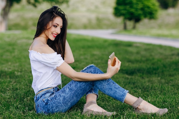 Chica joven sentada en un parque haciéndose una autofoto mientras sonrie y se toca el pelo