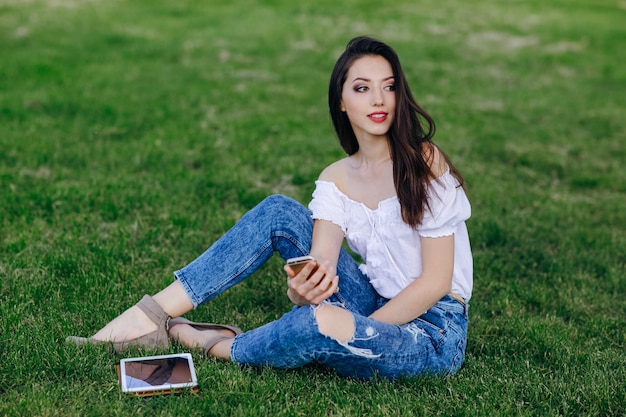 Chica joven sentada en un parque con una tablet en el césped y un móvil en la mano
