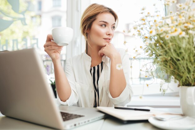 Chica joven sentada en el restaurante con una taza en la mano y una computadora portátil en la mesa. Retrato de una chica hermosa con cabello rubio descansando su barbilla en la mano mirando cuidadosamente a un lado en el café