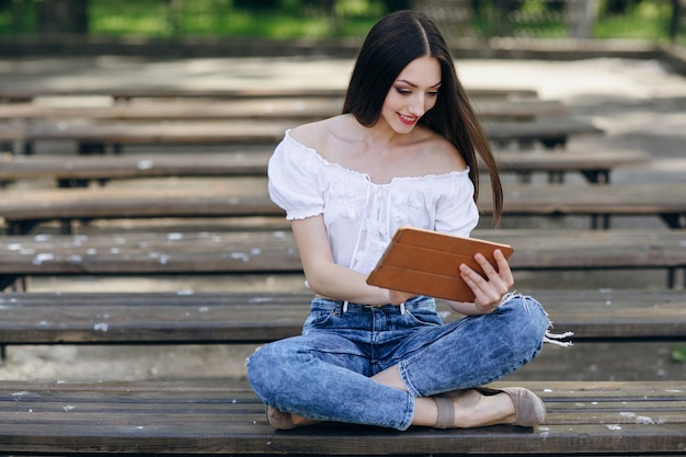 Chica jóven sonriendo y sentada en un banco de madera con una tablet