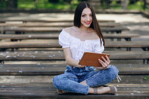 Chica jóven sonriendo y sentada en un banco de madera con una tablet