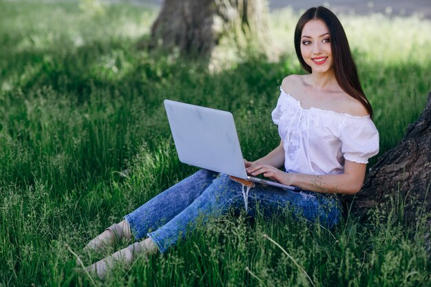 Chica jóven sonriendo sentada en el césped con un portátil en las piernas