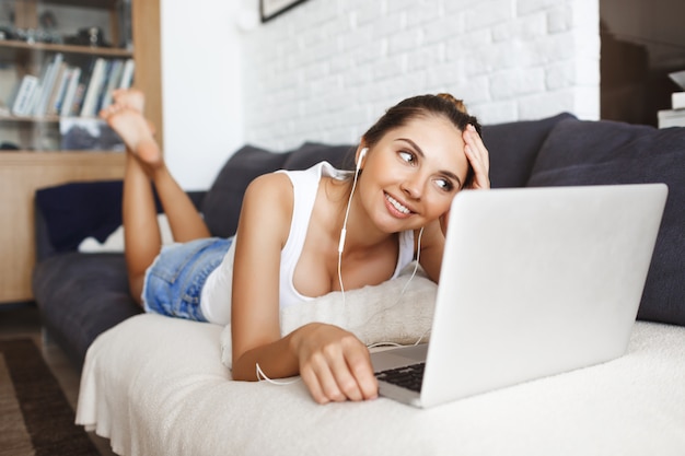 Chica joven sonriente atractiva que pone en el sofá en la sala de estar con la computadora portátil.