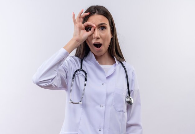 Chica joven sorprendida con bata médica estetoscopio mostrando gesto de mirada sobre fondo blanco aislado