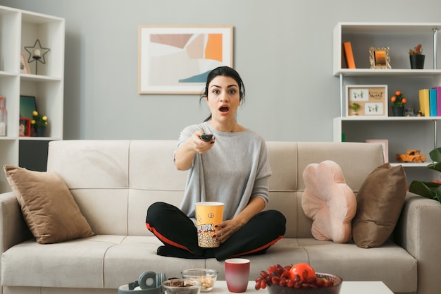 Chica joven sorprendida con un cubo de palomitas de maíz sosteniendo el control remoto del televisor, sentado en el sofá detrás de la mesa de café en la sala de estar