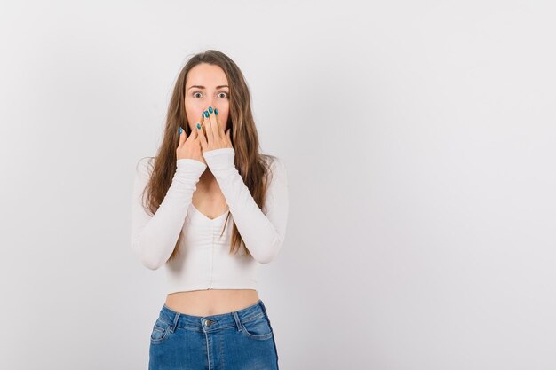 Chica joven sorprendida está cubriendo la boca con las manos sobre fondo blanco.