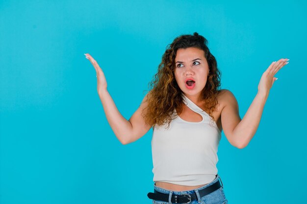 Chica joven sorprendida está mirando hacia arriba levantando las manos sobre fondo azul.