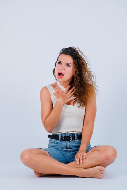 Chica joven sorprendida está mirando hacia arriba sentándose en el suelo sobre fondo blanco.