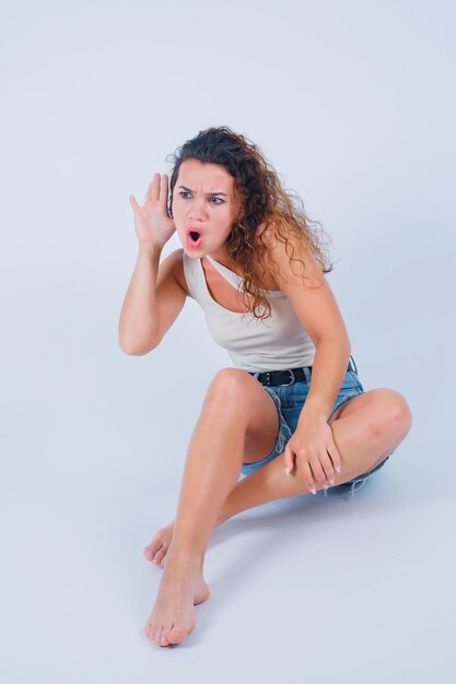 Chica joven sorprendida está sosteniendo la mano detrás de la oreja sentándose en el suelo sobre fondo blanco.