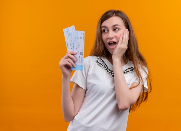 Chica joven sorprendida que sostiene los billetes de avión que pone la mano en la mejilla en la pared anaranjada aislada con el espacio de la copia