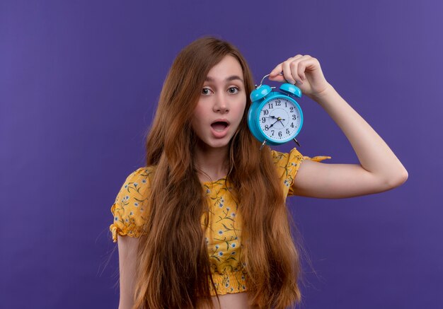 Chica joven sorprendida que sostiene el reloj de alarma en la pared púrpura aislada