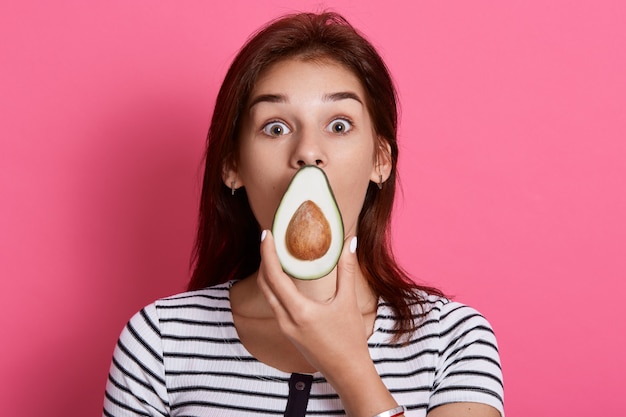 Chica joven sorprendida en ropa casual que cubre la boca con fruta fresca de aguacate verde madura aislada en la pared rosa, con camiseta a rayas, se encuentra con ojos grandes.