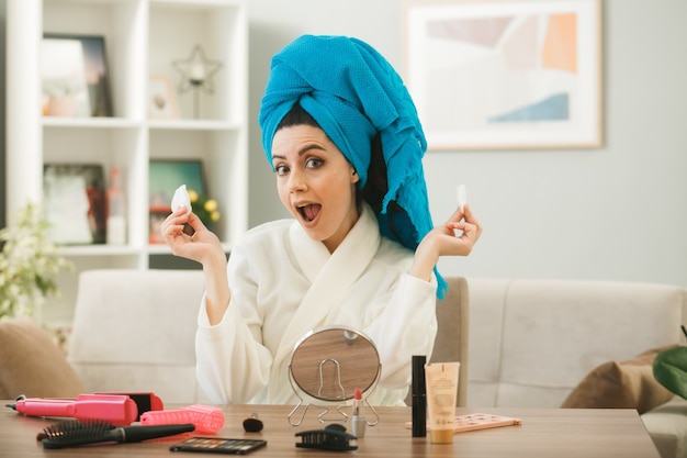 Chica joven sorprendida sosteniendo una esponja sentada a la mesa con herramientas de maquillaje en la sala de estar